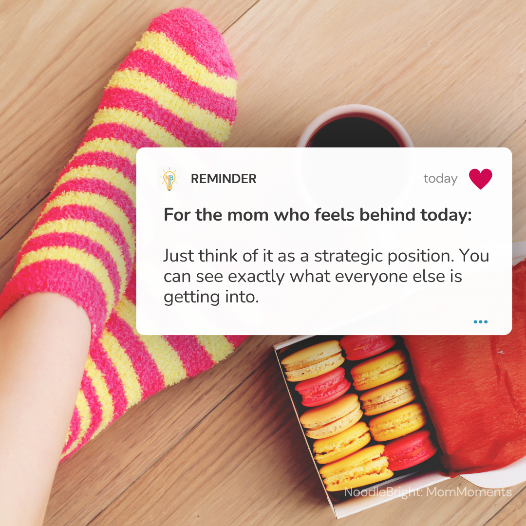A person's foot wearing a bright pink and yellow striped fuzzy sock on a wooden floor, next to a cup of coffee and an open box of macarons. A white text overlay reads: 'REMINDER For the mom who feels behind today: Just think of it as a strategic position.