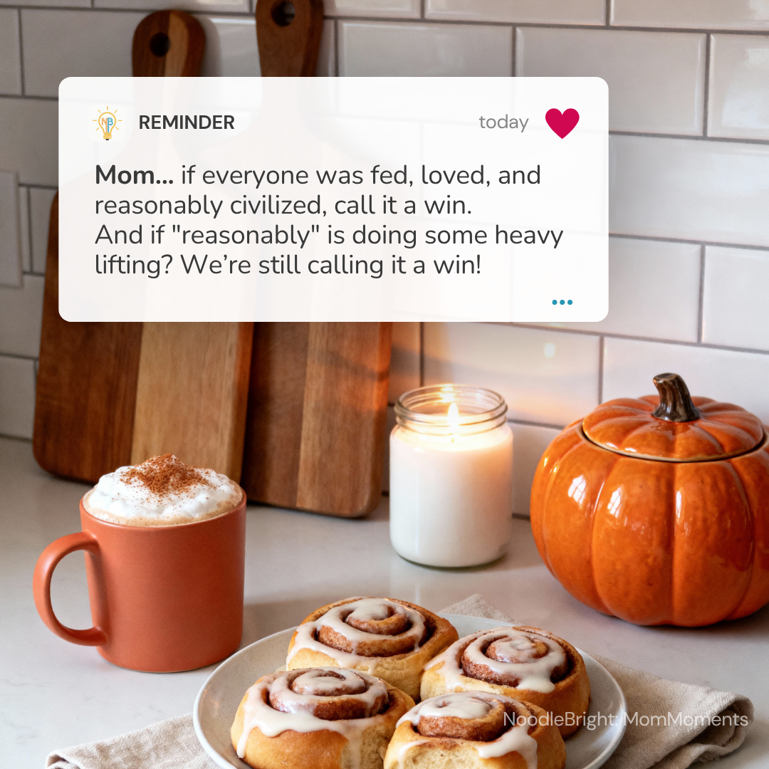 A cozy kitchen scene with a white text reminder card overlaid at the top, showing a warm mug topped with whipped cream and cinnamon, a plate of cinnamon rolls, a lit candle, and an orange pumpkin against a white subway tile backsplash.