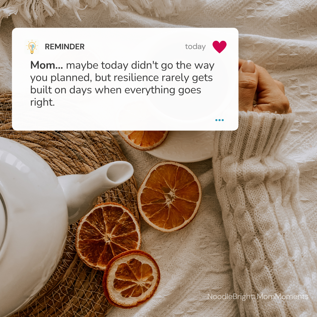 A warm lifestyle scene with a white teapot, dried orange slices, and a knitted cream sweater on a beige fabric surface. A white reminder card appears over the upper left, reading a message to a mom about resilience, with a small 'NoodleBright Mom Moments' watermark in the lower right corner.