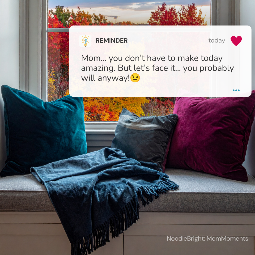 A cozy window seat with four decorative throw pillows in teal, dark gray, and magenta, draped with a dark blue fringed blanket. A large window behind the seat shows a colorful autumn tree view.