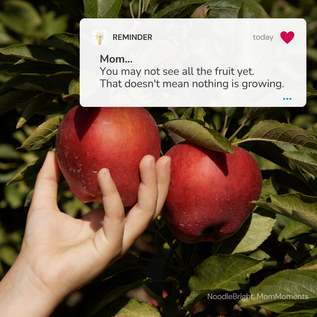 A hand holds a ripe red apple hanging from a leafy tree branch, with another red apple beside it. A white text overlay says, 'Mom.